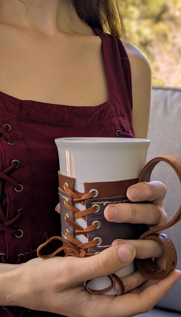 Hands closed around a a hot mug of coffee, fingers protected form burning by a brown and purple leather sleeve with adjustable laces and a handle.