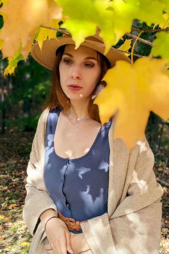 Portrait of writer and artist Amanda Surowitz looking off to the side with a relaxed expression while standing under a maple tree just starting to turn yellow in autumn.