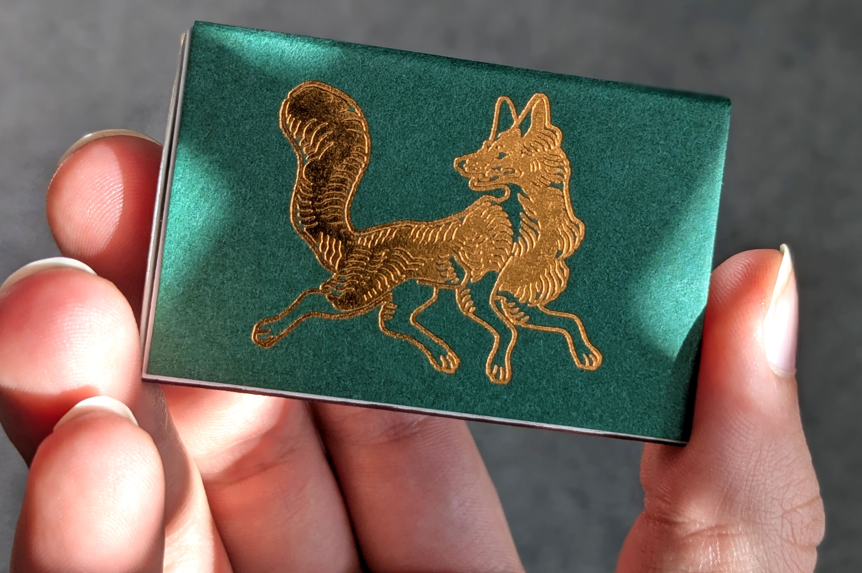 A green matchbox from Cantrip Candles with a metallic medieval illustration of a fox.