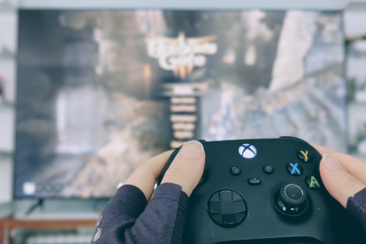 Hands holding up an Xbox controller in front of an out of focus screen displaying the pause menu for Baldur's Gate 3.